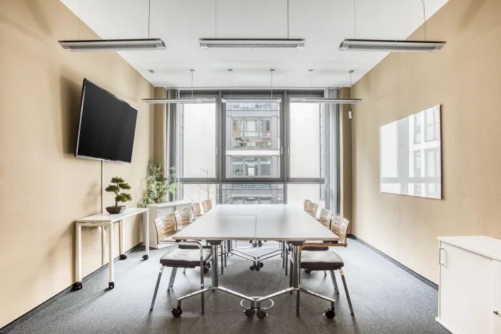 Heller Besprechungsraum mit Bildschirm, Whiteboard, Tisch und Stühlen, sowie Bodentiefe Fenster